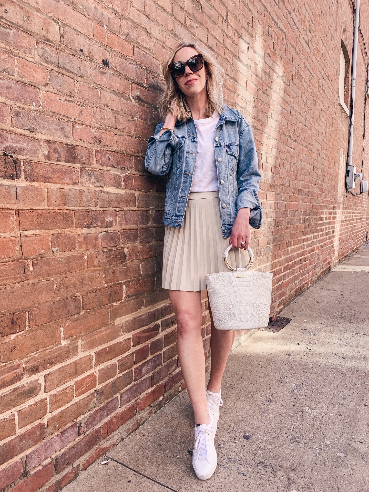 https://meagansmoda.com/wp-content/uploads/2020/03/Meagan-Brandon-fashion-blogger-of-Meagans-Moda-wears-denim-jacket-with-beige-faux-leather-pleated-skirt-and-white-sneakers.jpg